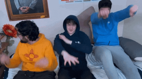 three young men are sitting on a bed with one wearing a yellow sweatshirt with a sun on it