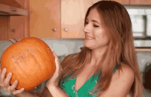 a woman in a green dress is holding a pumpkin in her hands in a kitchen .