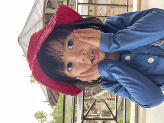 a little girl wearing a red hat and a blue shirt has her hands on her chin