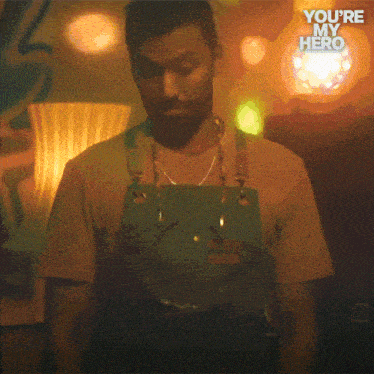 a man wearing an apron is standing in a dark room with the words " you 're my hero " written above him