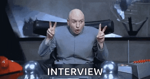 dr. evil is giving a peace sign while sitting at a desk with the word interview written on it .