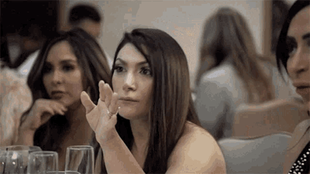 a group of women are sitting at a table with wine glasses and a woman is making a gesture with her hand .