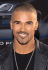 a man with a shaved head and a beard is smiling while wearing a leather jacket and a necklace .