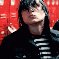 a young man wearing a striped shirt and a black jacket is smoking a cigarette in front of red lockers