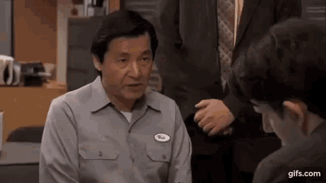 a man in a gray shirt with a name tag on his chest is talking to another man in a suit .