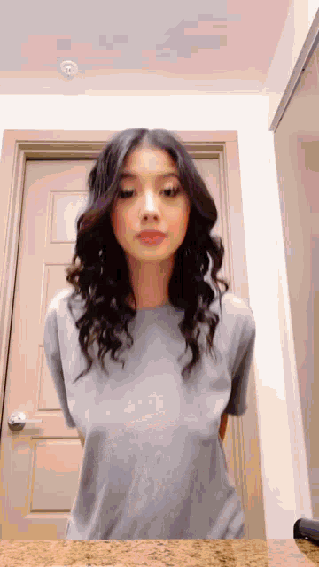 a woman in a grey shirt is standing in front of a door and looking at the camera