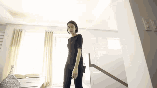 a woman in a black jumpsuit is standing in a room next to a window