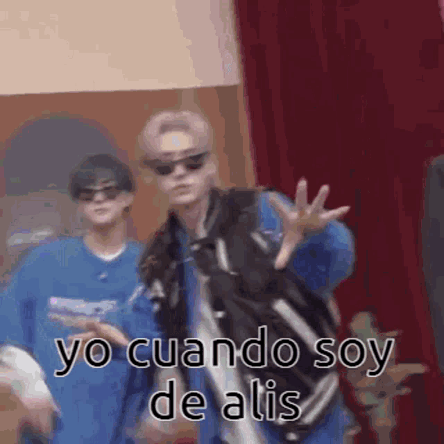 a man wearing sunglasses is dancing in front of a red curtain while another man watches .
