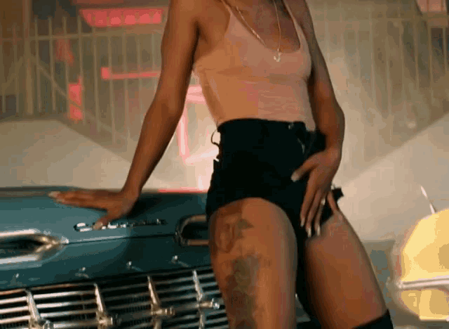 a woman in shorts and a tank top stands next to a blue car