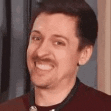 a man with a mustache is smiling and wearing a red shirt .