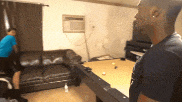 two men playing pool in a living room with an air conditioner on the wall above the pool table