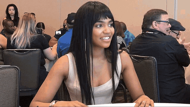 a woman in a white tank top sits at a table in a crowd