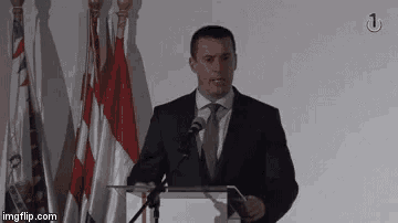 a man in a suit and tie is giving a speech at a podium with flags in the background