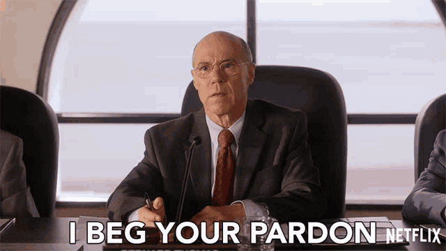 a man in a suit and tie is sitting at a table and says i beg your pardon