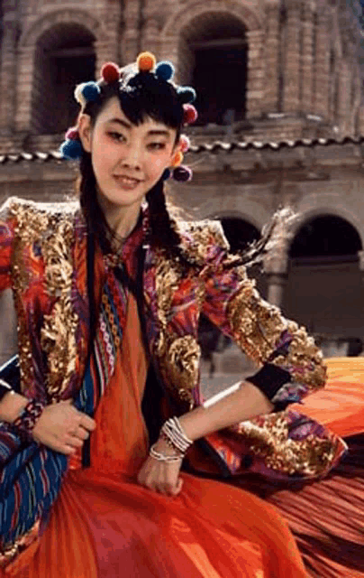 a woman wearing a colorful jacket and a crown of pom poms is sitting in front of a building .
