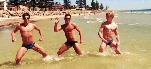 three shirtless men in swim trunks are dancing in the ocean .