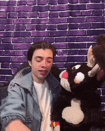 a man is sitting in front of a purple brick wall holding a stuffed animal .