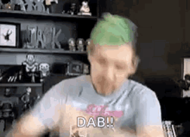 a man with green hair is sitting in front of a shelf and making a face .
