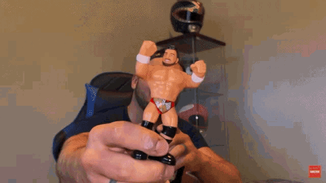 a man is holding a toy wrestler in his hand with a subscribe button on the bottom right