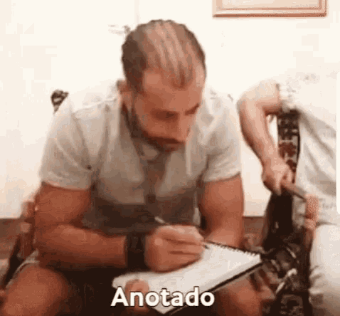 a man is sitting on a couch writing in a notebook with the word anotado written next to him .