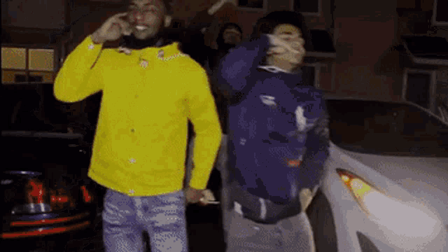a man in a yellow jacket is talking on a cell phone next to another man in a purple jacket .