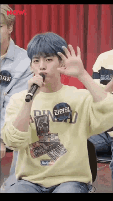 a man with blue hair is holding a microphone and wearing a yellow shirt that says ' now ' on it