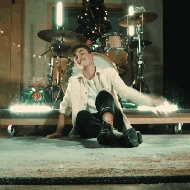a young man is sitting on the floor in front of a drum set and a christmas tree