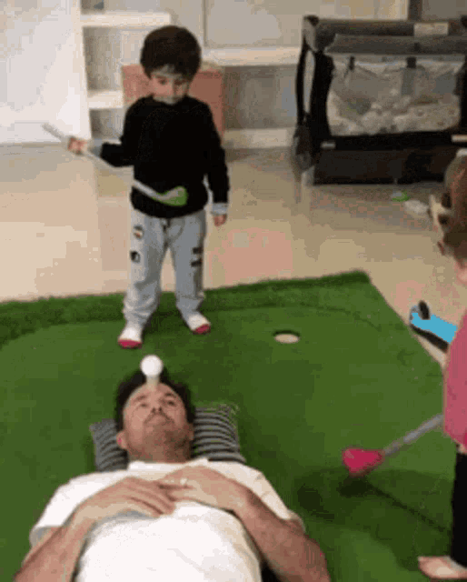 a man is laying on a pillow playing golf with two children standing around him