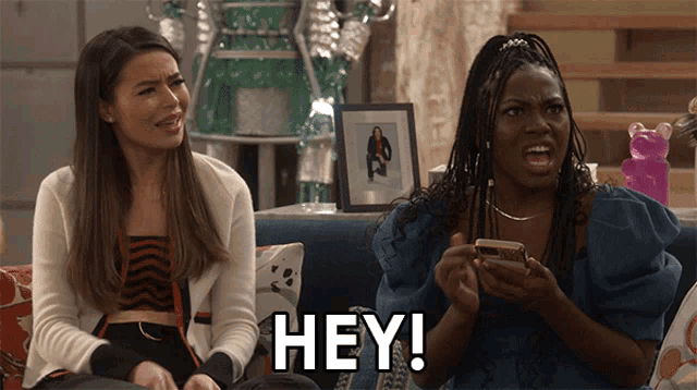 two women are sitting on a couch and one of them is saying " hey "
