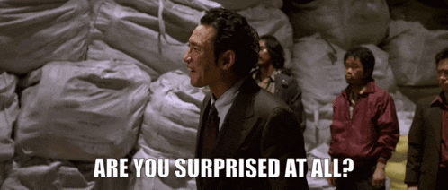 a man in a suit and tie stands in front of a pile of bags and says are you surprised at all