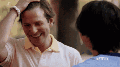 a man in a pink polo shirt is smiling and touching his hair while talking to another man .