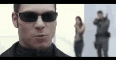 a man wearing sunglasses is standing in front of a woman holding a gun in a room .