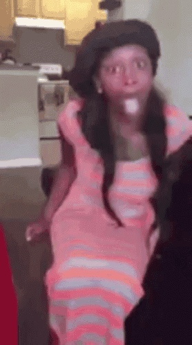 a girl in a pink dress and hat is standing in a kitchen .
