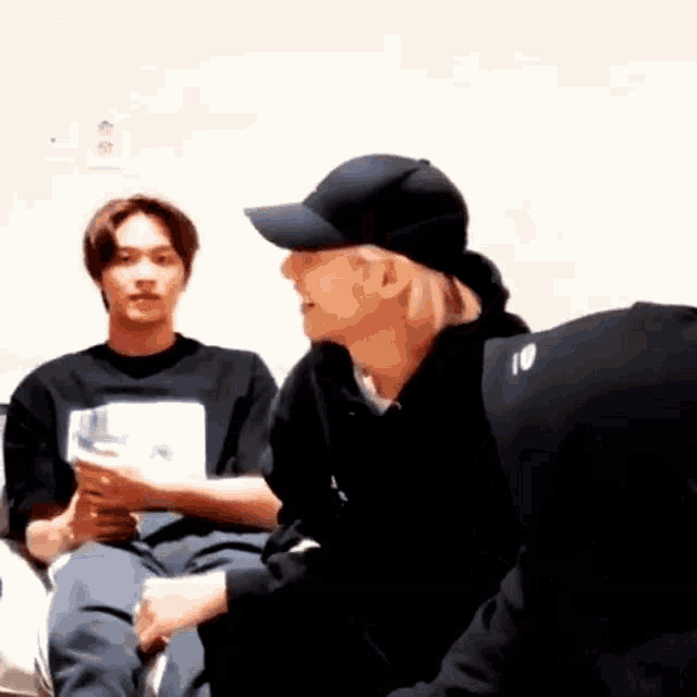 a couple of men are sitting next to each other in a room . one of the men is wearing a baseball cap .