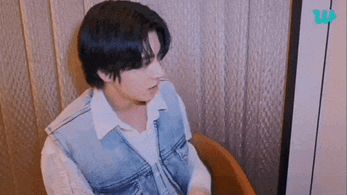 a young man is sitting in a chair wearing a denim vest .