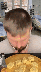 a man with a beard is eating noodles from a cutting board in a hospital room .