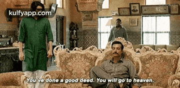 a man is sitting on a couch in a living room with a man standing behind him .