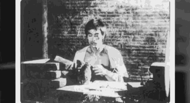 a black and white photo of a man sitting at a desk with a brick wall in the background .