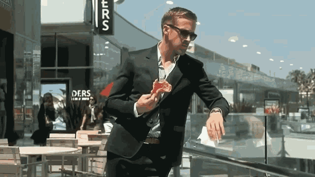a man in a suit eating a slice of pizza in front of a ders sign