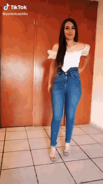 a woman in a white off the shoulder top and blue jeans is standing in a room .