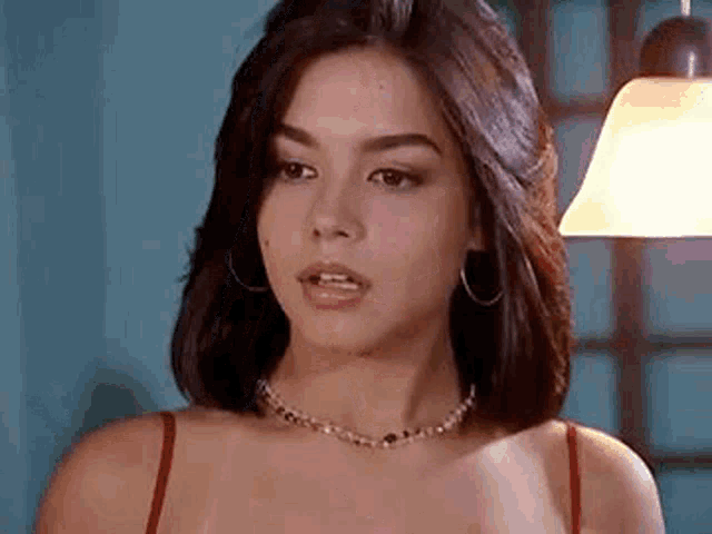 a woman wearing a red tank top and a pearl necklace is standing in front of a lamp .
