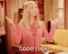 a woman in a pink cardigan is standing in a living room with her arms outstretched and says `` good luck '' .