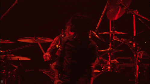 a man playing a guitar in front of a drum set with a red light behind him