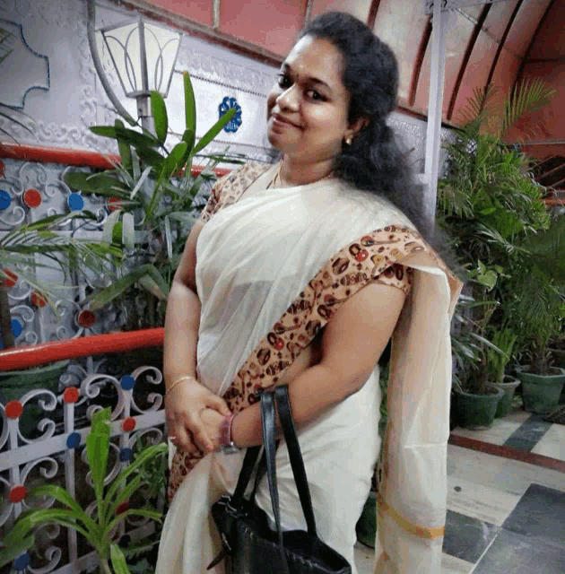 a woman in a white saree with a black purse stands in front of potted plants