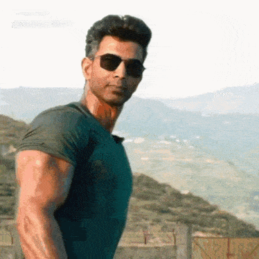 a man wearing sunglasses and a green shirt stands in front of a mountain