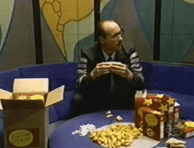 a man is sitting on a couch eating a sandwich with a box of chips in front of him