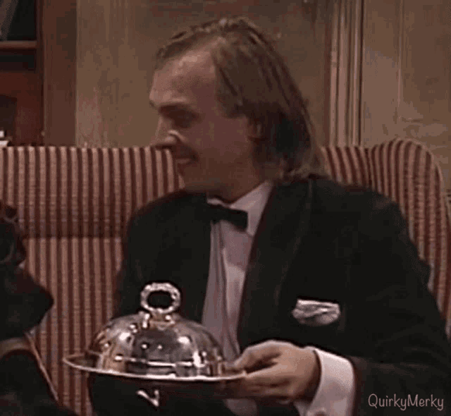 a man in a tuxedo is holding a silver tray with a dome lid .