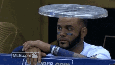 a man with a plastic lid on his head sits in a dugout with mlb.com in the corner