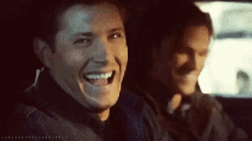 two men are laughing while sitting in a car and one of them is wearing a black jacket