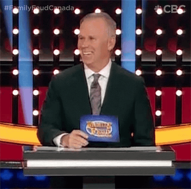 a man in a suit and tie is standing at a podium holding a sign that says family feud canada .
